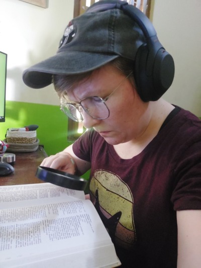 Eine weiße Person mit Brille, Kappe und Kopfhörern beugt sich mit einer Lupe in der Hand über ein großes aufgeschlagenes Wörterbuch.